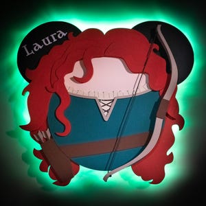 May include: A layered paper art piece featuring a cartoon character with red hair, a teal dress, and a bow and arrow. The name "Laura" is written on a black Mickey Mouse ear. The artwork is illuminated with a green glow.
