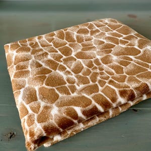 May include: A folded plush fabric featuring a repeating giraffe print pattern with irregular brown patches separated by thin white lines. The material has a soft, fuzzy texture and is arranged in a neat stack, showcasing the detailed animal-inspired design.