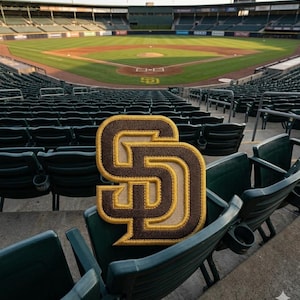 May include: An embroidered San Diego Padres logo, a brown and gold "SD", is shown in a baseball stadium. The stadium has green seating and a baseball field in the background.