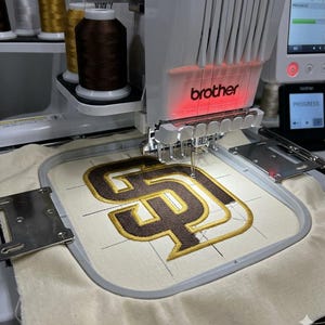 May include: An embroidery machine by Brother is shown in operation, stitching a logo onto a cream-colored fabric. The logo features a brown and gold design. Spools of thread in various colors are visible above the machine.