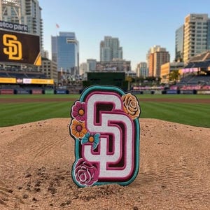 Puede incluir: Parche bordado con el logotipo de los Padres de San Diego en blanco y rosa, contorneado en turquesa y marrón. El parche está decorado con acentos florales coloridos, incluyendo flores rosas, naranjas y amarillas. El parche está colocado en un campo de béisbol.