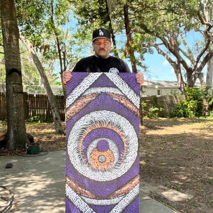 May include: A rectangular purple artwork with a circular design in the center. The artwork features concentric circles in white, orange, and brown, with a textured background. The artist is holding the artwork outdoors.