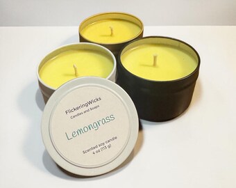 May include: Four round, scented soy candles in metal tins. The candles have a bright yellow wax and white wicks. One lid is open, revealing the text "Lemongrass" and "Scented soy candle 4 oz (113 g)". The tins are in various colors.
