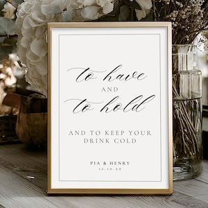 May include: A framed sign with gold trim displays the text "To have and to hold" in elegant script. Below, it reads "And to keep your drink cold." The sign includes names and a date. The sign is on a wooden surface, with flowers in the background.