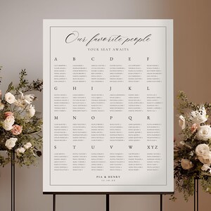 Puede incluir: Un plano de asientos de boda con el texto "Our favorite people, Your seat awaits." El gráfico está sobre un soporte negro, rodeado de arreglos florales y velas. El gráfico enumera los nombres alfabéticamente.