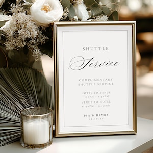 May include: A gold-framed sign with the words "SHUTTLE Service" in elegant script. The sign includes details about a complimentary shuttle service, with a candle in a gold-rimmed glass and a floral arrangement in the background. Wedding decor.
