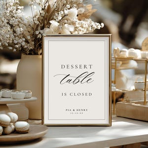 May include: A framed sign with the text "DESSERT table IS CLOSED" in elegant script, with the names "PIA & HENRY" and a date below. The sign is set on a table with desserts and floral arrangements.