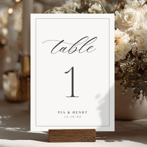 May include: A table number card with the word "table" in elegant script above the number "1". The card includes the names "PIA & HENRY" and a date. The card is displayed in a wooden holder, with a floral arrangement in the background.