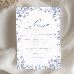 以下が含まれることがあります： 青い花の装飾と「Louisa」の名前が書かれた白い招待状。カードには、「私の結婚式で、私の何か青いものになってくれませんか？」という心からのメッセージが書かれています。愛を込めて、クリスティーナ。