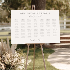 May include: A cream-colored wedding seating chart on a wooden easel. The sign reads "OUR FAVORITE PEOPLE find your seat" in elegant black script. Below, names are listed under table numbers. The names are in a simple, easy-to-read font. The names are listed under table numbers.
