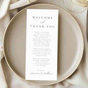 May include: A wedding reception table setting with a white card that reads "WELCOME and THANK YOU" in elegant script. The card is on a beige plate, with a knife and a glass of wine visible. Delicate white flowers are in the background.