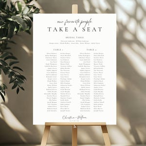 Op de afbeelding: Een trouwzittingsoverzicht op een houten ezel. Het bord leest "our favorite people TAKE A SEAT" in elegant schrift. Namen staan vermeld onder "Bridal Table", "Table 1" en "Table 2". De namen zijn in zwarte letters op een crèmekleurige achtergrond.