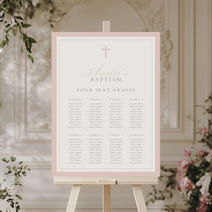 May include: A rectangular baptism seating chart sign with a dusty pink border and a light beige center. The sign features a cross icon, the text Amelia's BAPTISM, YOUR SEAT AWAITS, and a list of eight tables with names, all displayed on a wooden easel.