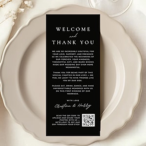 May include: A black welcome and thank you card with white text, placed on a white plate. The card includes a message of gratitude, names, and a QR code. The card is a wedding favor.