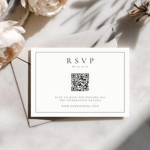 May include: A white RSVP card with a black border, featuring the word "RSVP" at the top, a QR code, and the text "Scan to RSVP and explore all the celebration details." The card is on a gray envelope, with white flowers in the background.