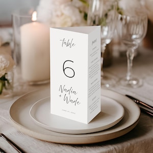 May include: A white, triangular table card with the text "table 6 Nadia + Wade June 27, 20XX" in black script. The card is on a white plate, with a fork and knife in the foreground. A lit candle and flowers are in the background.