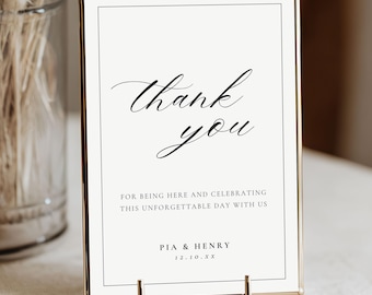 May include: A framed sign with the words "Thank you" in elegant script. Below, text reads "For being here and celebrating this unforgettable day with us." The sign is held by a gold stand, perfect for a wedding or event.