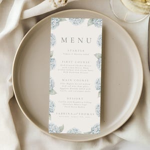 May include: A wedding menu card with a floral design featuring blue hydrangeas. The menu lists the courses: Starter, First Course, Main Course, and Dessert. The names Sabrina and Thomas are at the bottom, along with the date.