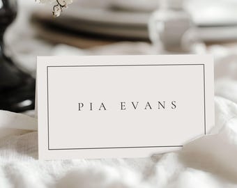May include: A rectangular place card with the name "PIA EVANS" printed in black, centered within a black border. The card sits on a white tablecloth, with blurred floral arrangements and tableware in the background.