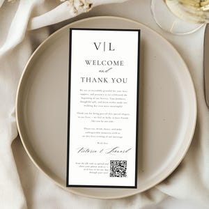 May include: A wedding reception place setting with a white plate, silverware, and a welcome card. The card has a black border and text that reads "WELCOME and THANK YOU" with the names "Victoria & Leonard" and a QR code.