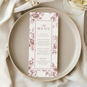 May include: A rectangular menu card with a floral design in shades of mauve and white, featuring the text "The Menu." The menu is placed on a white plate, with a knife and a glass of wine visible in the background. The menu lists the courses: Starter, First Course, Main Course, and Dessert.