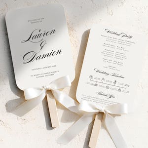 May include: Two white wedding fans with black text and a cream-colored ribbon. One fan reads "Welcome to the Wedding of Lauren & Damien" and the other lists the wedding party and timeline. The fans have wooden handles.