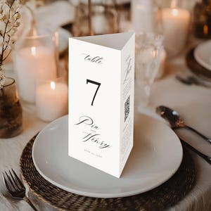 Op de afbeelding: Een driehoekige tafelnummerkaart met het zwarte nummer 7 en de namen "Pia and Henry" in sierlijke letters. De kaart staat op een witte plaat, met een geweven placemat en bestek.