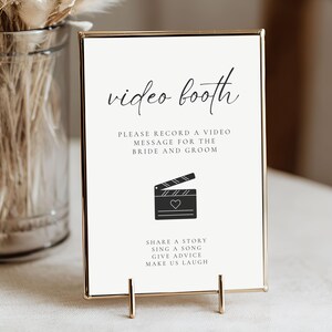 May include: A framed sign with the words "video booth" in elegant script. The sign includes instructions to record a video message for the bride and groom, along with a film clapper icon and a list of suggestions.
