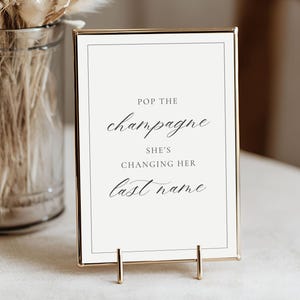 Könnte beinhalten: Ein rechteckiges Schild mit dem Text "Pop the Champagne, She's Changing Her Last Name" in eleganter Schrift. Das Schild ist in einem goldenen Rahmen und steht auf einem goldenen Ständer.