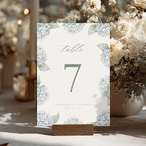 May include: A table number card with a floral design. The card features a light beige background with blue hydrangea flowers and green leaves. The word "table" and the number "7" are displayed in green. The names "Sabrina and Thomas" are also printed on the card.