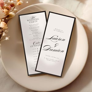 May include: Two wedding programs with black borders and white backgrounds are displayed on a cream-colored plate. The programs feature elegant black text, including the names "Lauren & Damien" and the wedding details. Dried flowers are in the background.