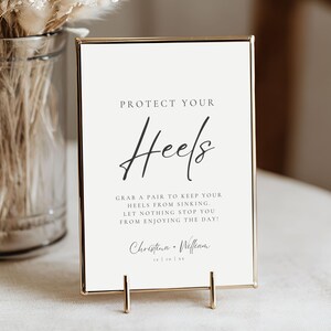 Puede incluir: Un letrero enmarcado con el texto "PROTECT YOUR Heels" en una elegante escritura. El letrero también incluye el texto "GRAB A PAIR TO KEEP YOUR HEELS FROM SINKING. LET NOTHING STOP YOU FROM ENJOYING THE DAY!" y los nombres "Christina + William".