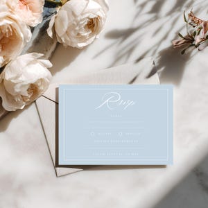 May include: A light blue RSVP card with white text and a white border. The card includes fields for names, acceptance or decline, and dietary requirements. The card is next to a light gray envelope and white and pink flowers.