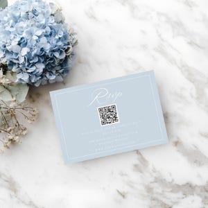 May include: A light blue RSVP card with white text and a QR code. The card is rectangular with a white border and is placed on a marble surface next to a blue hydrangea flower.