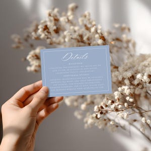 May include: A light blue wedding details card with white text and a white border. The card reads "Details" at the top, followed by information about the reception, additional details, and a website address. The card is held in front of a blurred floral background.