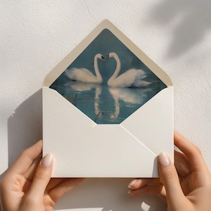 May include: A white envelope with a watercolor painting of two swans forming a heart shape with their necks. The swans are reflected in the water. The envelope is held open, revealing the artwork inside.