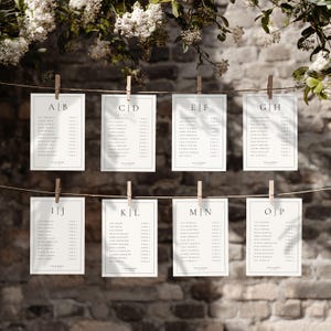 Op de afbeelding: Acht witte rechthoekige trouwzittingkaarten hangen met houten wasknijpers aan een touwtje. Elke kaart heeft een zwarte rand en bevat gastennamen en tafelindelingen. De kaarten staan tegen een bakstenen muur, met bloemaccenten.