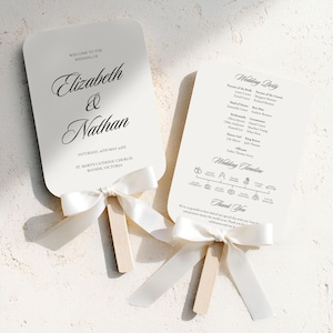 May include: Two white wedding fans with text, tied with white satin ribbons. One fan reads "Elizabeth & Nathan" and the wedding date. The other fan lists the wedding party and timeline. The fans have wooden handles.