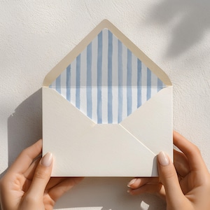 May include: An open cream-coloured envelope featuring a decorative interior lining with vertical light blue watercolour stripes. The envelope has a pointed flap and a textured paper finish, displaying a clean and simple design for stationery.
