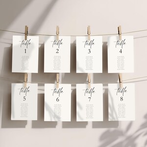 May include: Eight white rectangular cards with the word "table" in elegant script, numbered 1-8, and lists of names. The cards are suspended by wooden clothespins on a string against a neutral background.
