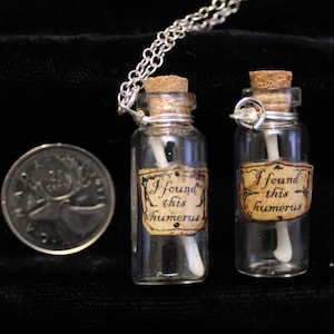 Könnte beinhalten: Zwei kleine Glasflaschen mit Korkstopfen und silbernen Ketten, jede mit einem kleinen Knochen und einem Etikett mit der Aufschrift "Ich habe diesen Humerus gefunden".