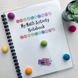 May include: A white notebook with a colorful watercolor design and the text "My Math Activity Notebook" and "EVERYDAY a little Grace". The notebook is closed and has a spiral binding. There are colorful felt balls scattered around the notebook.