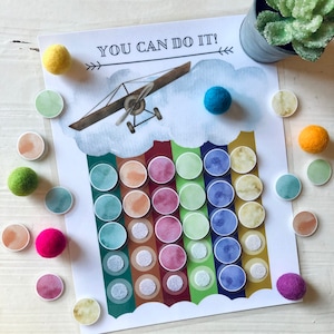 Puede incluir: Una hoja de actividad imprimible con el texto "You Can Do It!" y un avión de dibujos animados volando sobre un arcoíris con círculos coloridos. Los círculos están destinados a ser cubiertos con bolas de fieltro.