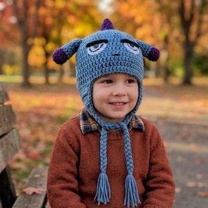 May include: A light blue crocheted hat featuring a purple top spike, two purple-tipped side flaps, and two braided chin straps with tassels. The hat has embroidered white and black eyes with a serious expression and a small stitched nose.