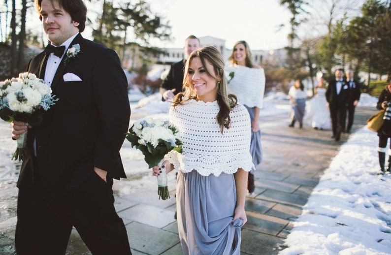 May include: A wedding party walks down a snow-covered path. The bride and bridesmaids wear gray dresses and white crocheted shawls. The groomsmen wear black tuxedos and carry bouquets of white flowers.