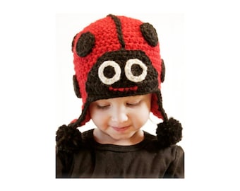 May include: A red and black crocheted hat designed to resemble a ladybug. The hat features a black face panel with two large white and black circular eyes, a small red stitched mouth, and black pom-poms hanging from the ear flaps.