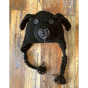 May include: Black crocheted dog hat with black buttons for eyes and a gray felt nose. The hat has long black braids with tassels.