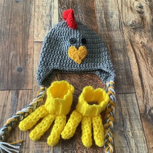 K&ouml;nnte beinhalten: Eine graue geh&auml;kelte H&uuml;hnerm&uuml;tze mit rotem Kamm und gelbem Schnabel. Die M&uuml;tze hat ein passendes Paar gelber geh&auml;kelter Fingerhandschuhe.