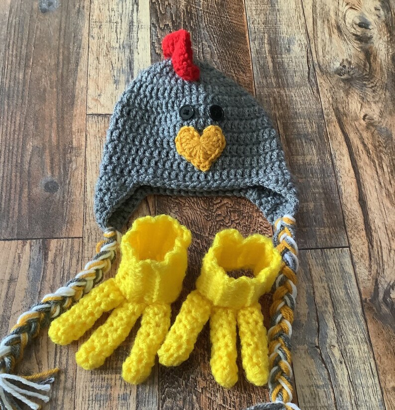 K&ouml;nnte beinhalten: Eine graue geh&auml;kelte H&uuml;hnerm&uuml;tze mit rotem Kamm und gelbem Schnabel. Die M&uuml;tze hat ein passendes Paar gelber geh&auml;kelter Fingerhandschuhe.