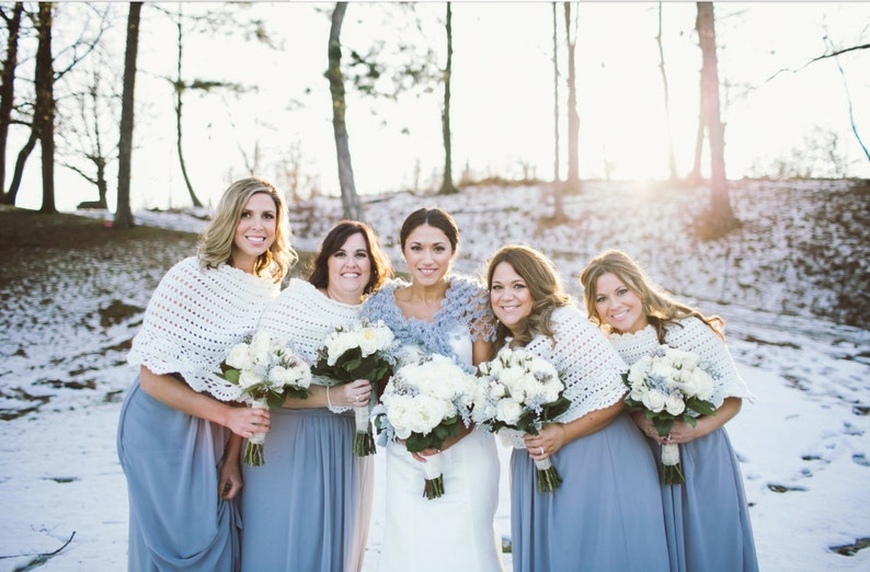May include: Five women in a snowy outdoor setting, wearing light blue dresses and white crocheted shawls. Each woman holds a bouquet of white flowers. The bride wears a white dress and a blue shawl.